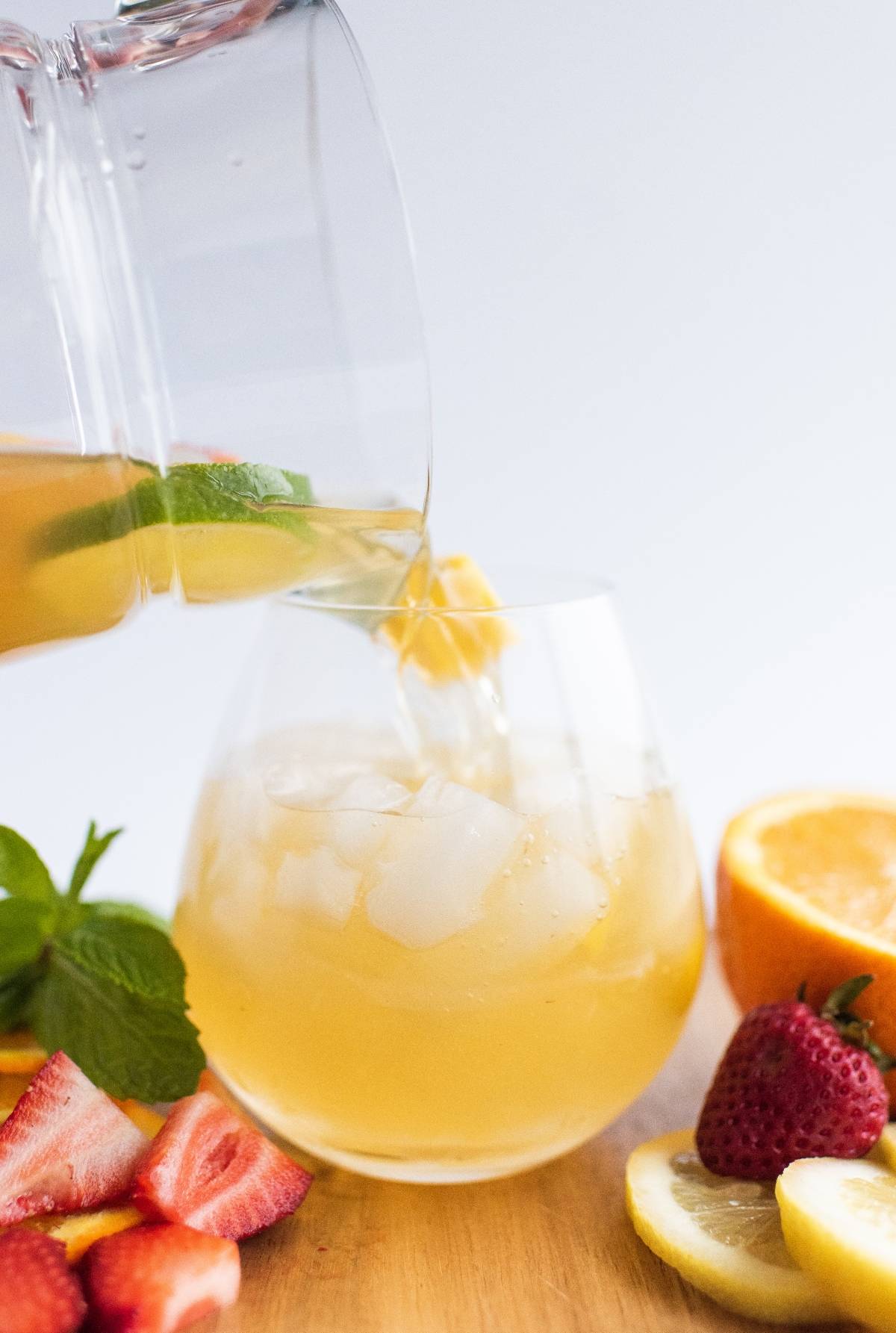 A pitcher pours iced tea into a glass with ice, surrounded by fresh strawberries, lemon, orange, and mint—perfect inspiration for a super easy sangria recipe or refreshing white sangria with summer fruit.