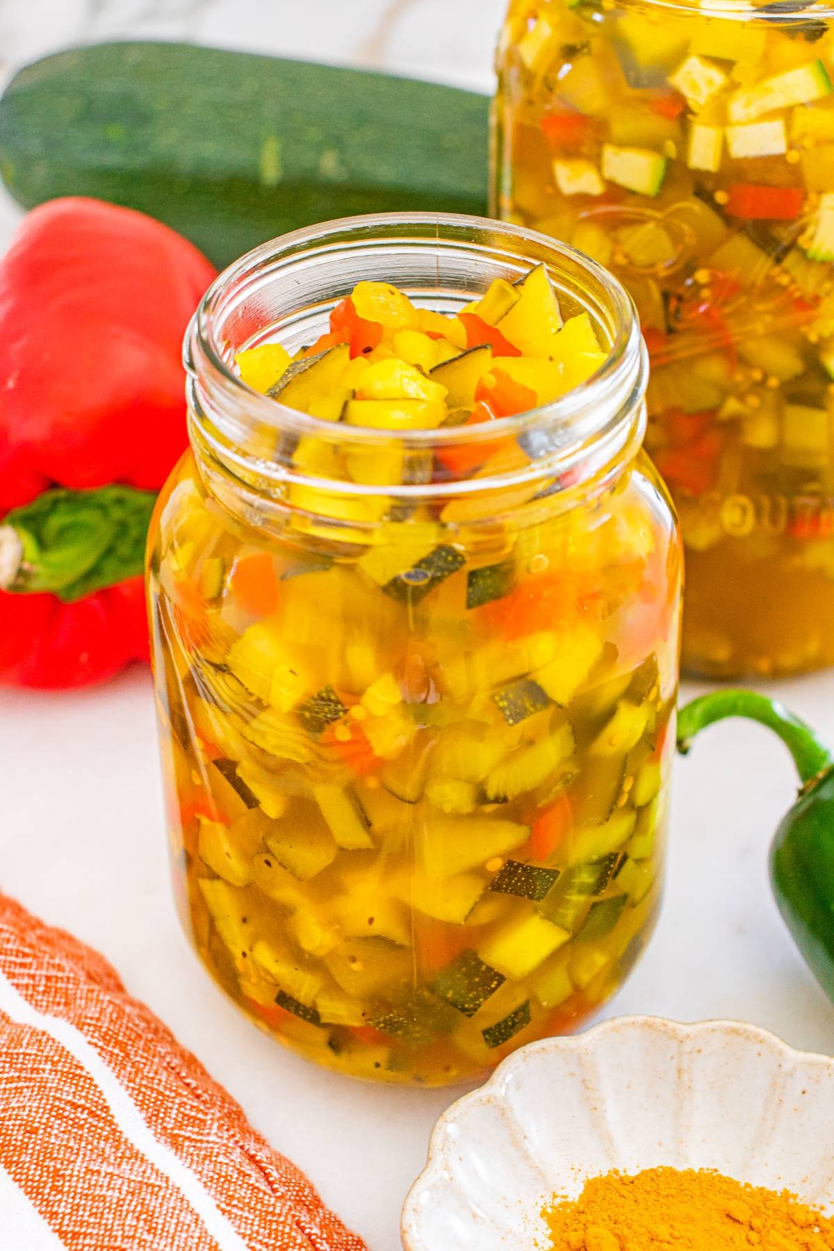 A jar of homemade zucchini relish with chopped vegetables, next to a red pepper and a cucumber.