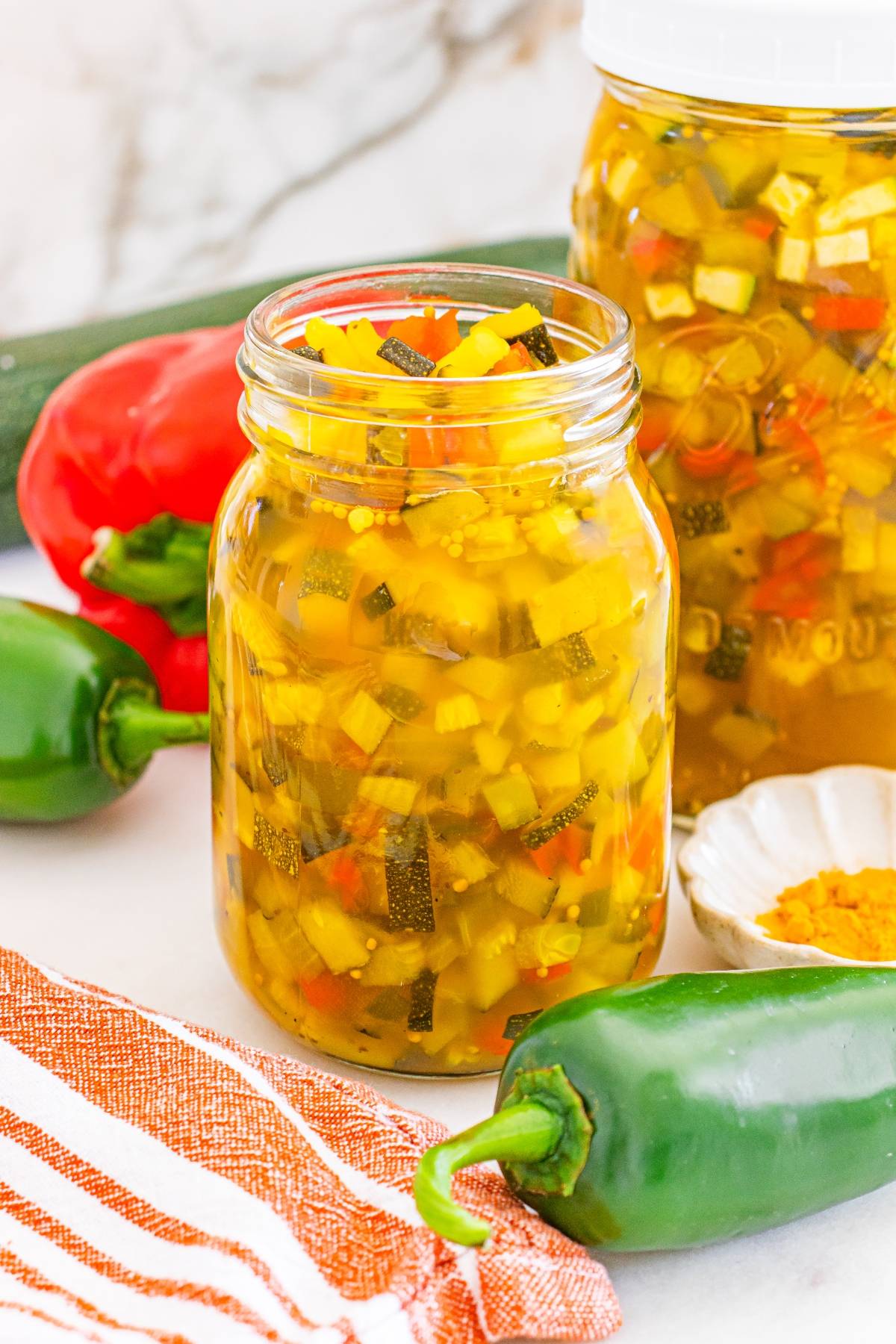 Open jar of homemade relish with diced vegetables, surrounded by fresh peppers and a striped towel.