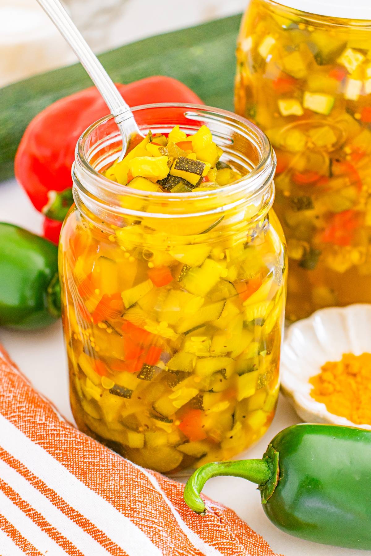 A jar of homemade relish with diced vegetables and a spoon, surrounded by fresh peppers and a striped towel.