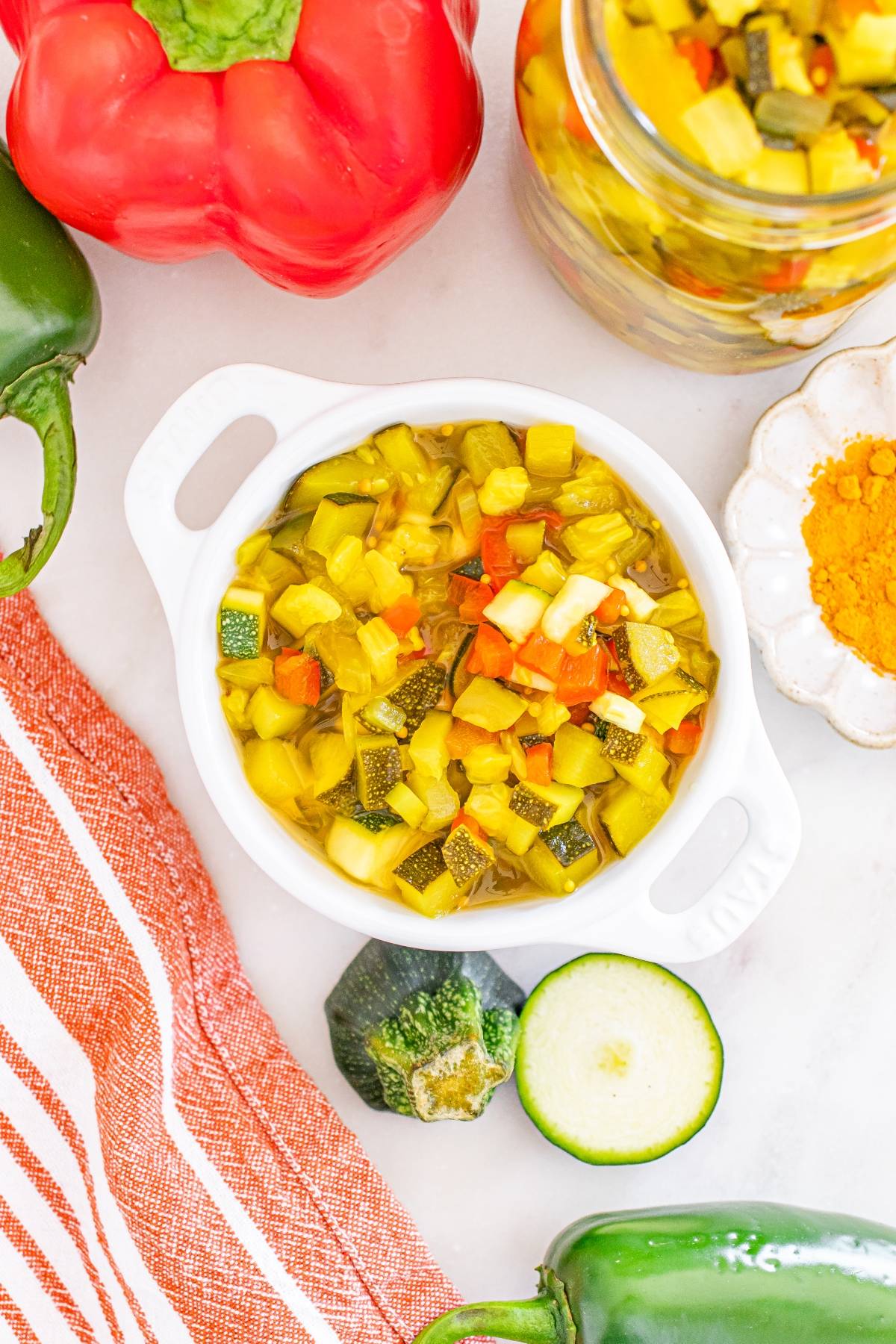 A bowl of colorful zucchini relish surrounded by fresh peppers, zucchini, a spice dish, and a red striped towel.