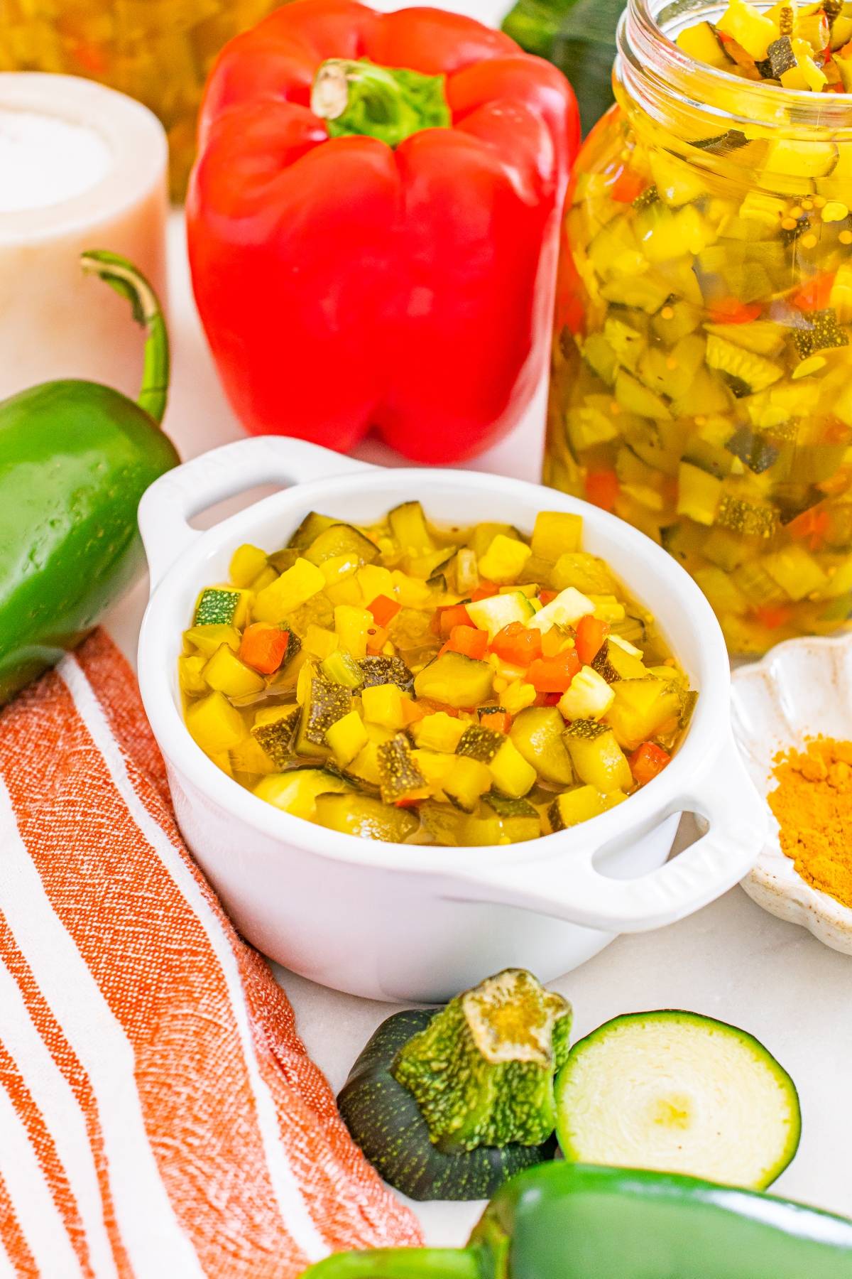 A bowl of chopped vegetable relish with jars, fresh vegetables, and a striped towel nearby.