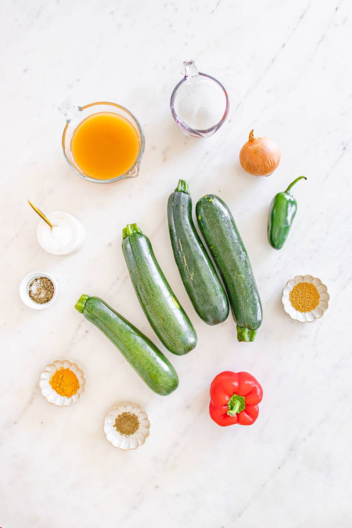 Three zucchinis, bell pepper, jalapeño, onion, broth, seasonings, and cream arranged on a white surface.