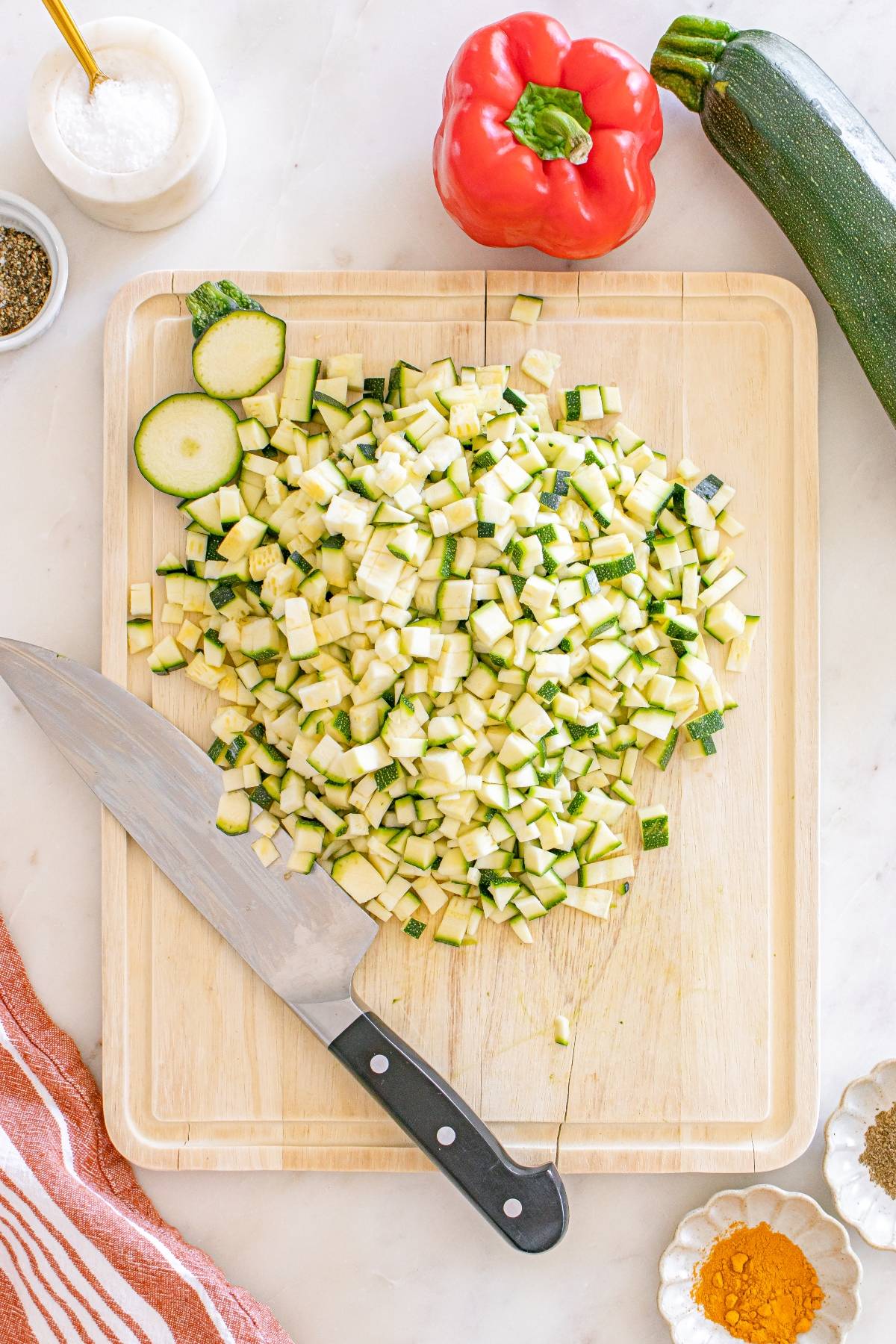 Diced zucchini on a cutting board with a knife, next to a red bell pepper and a whole zucchini.