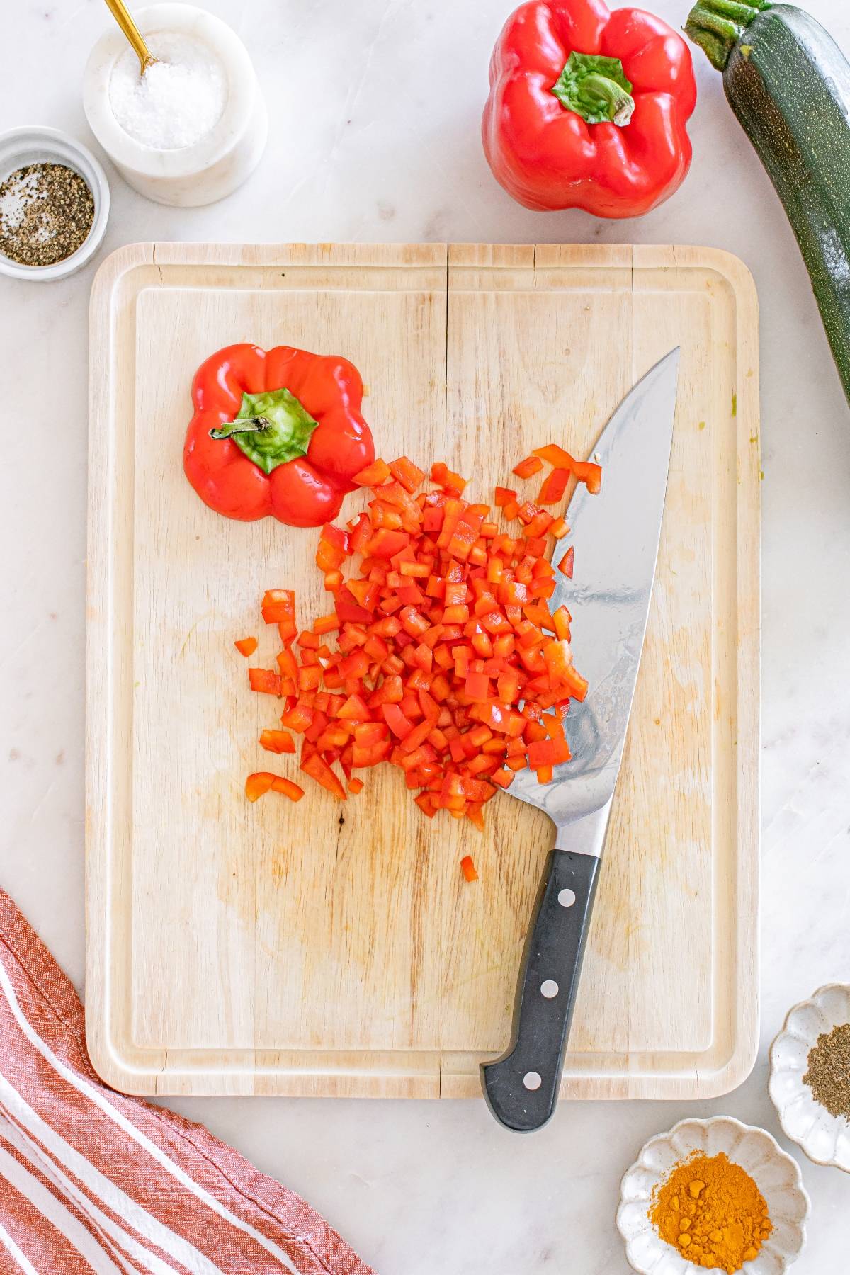 Chopped red bell pepper on a cutting board with a knife, whole pepper, zucchini, and spices nearby.