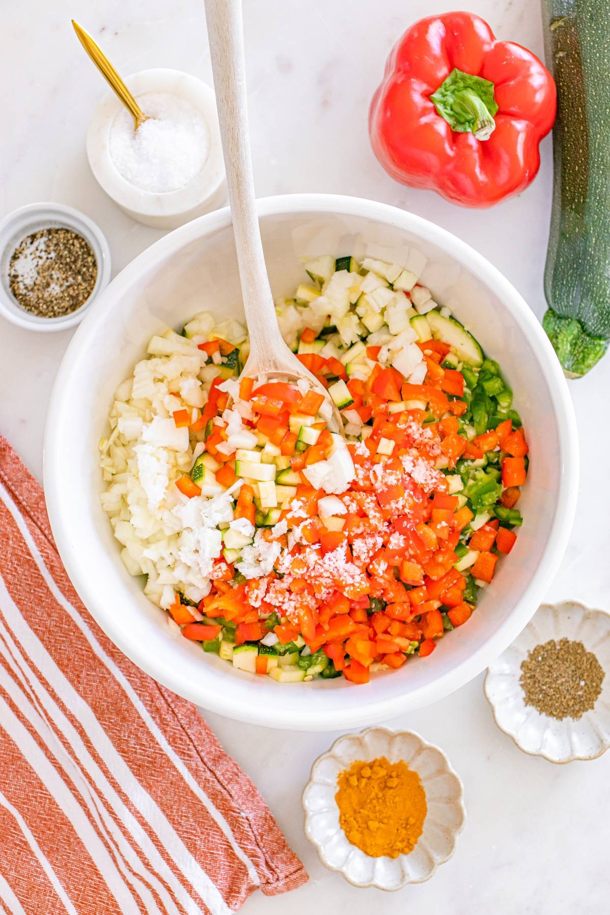 White bowl with chopped vegetables and a wooden spoon, surrounded by spices and a red bell pepper on a countertop.