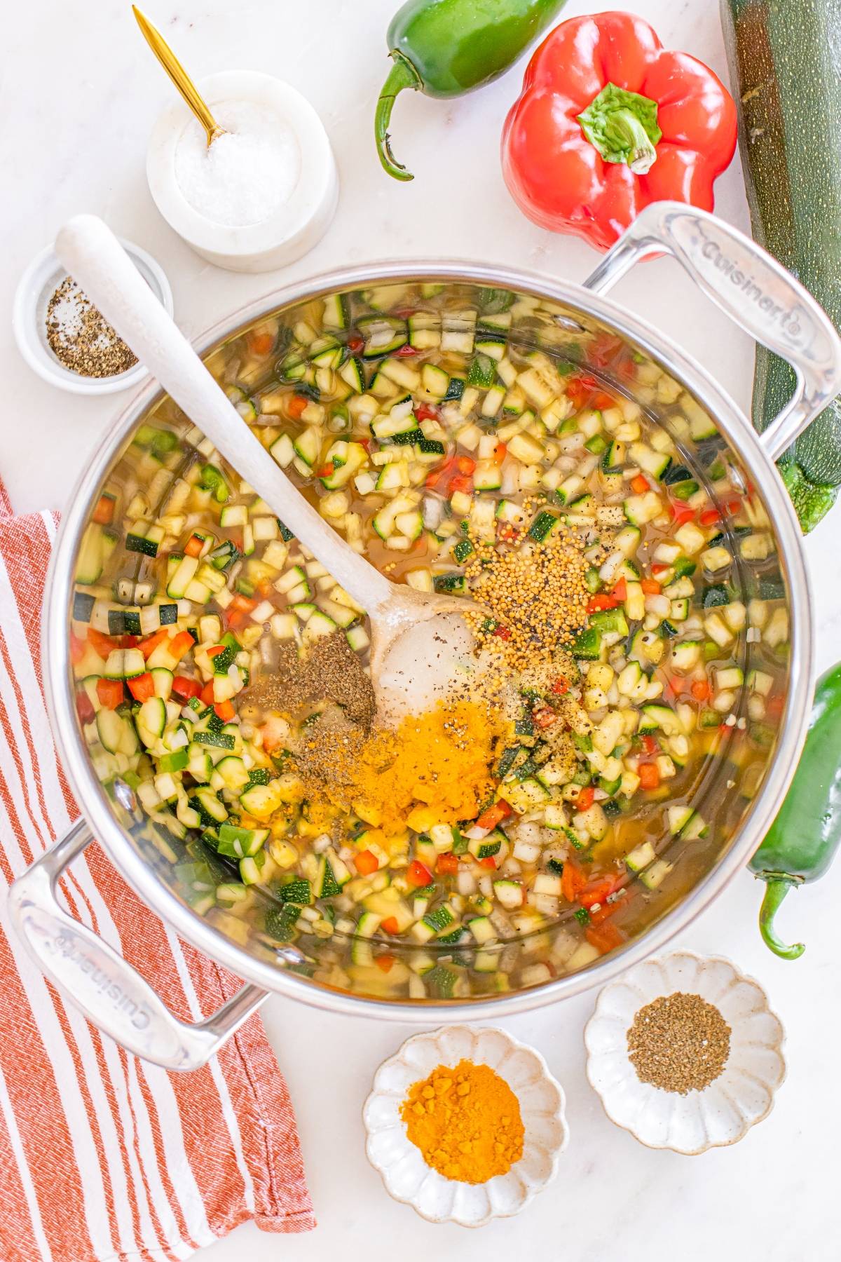 Chopped vegetables and spices in a pot with a wooden spoon, surrounded by fresh ingredients and seasonings.