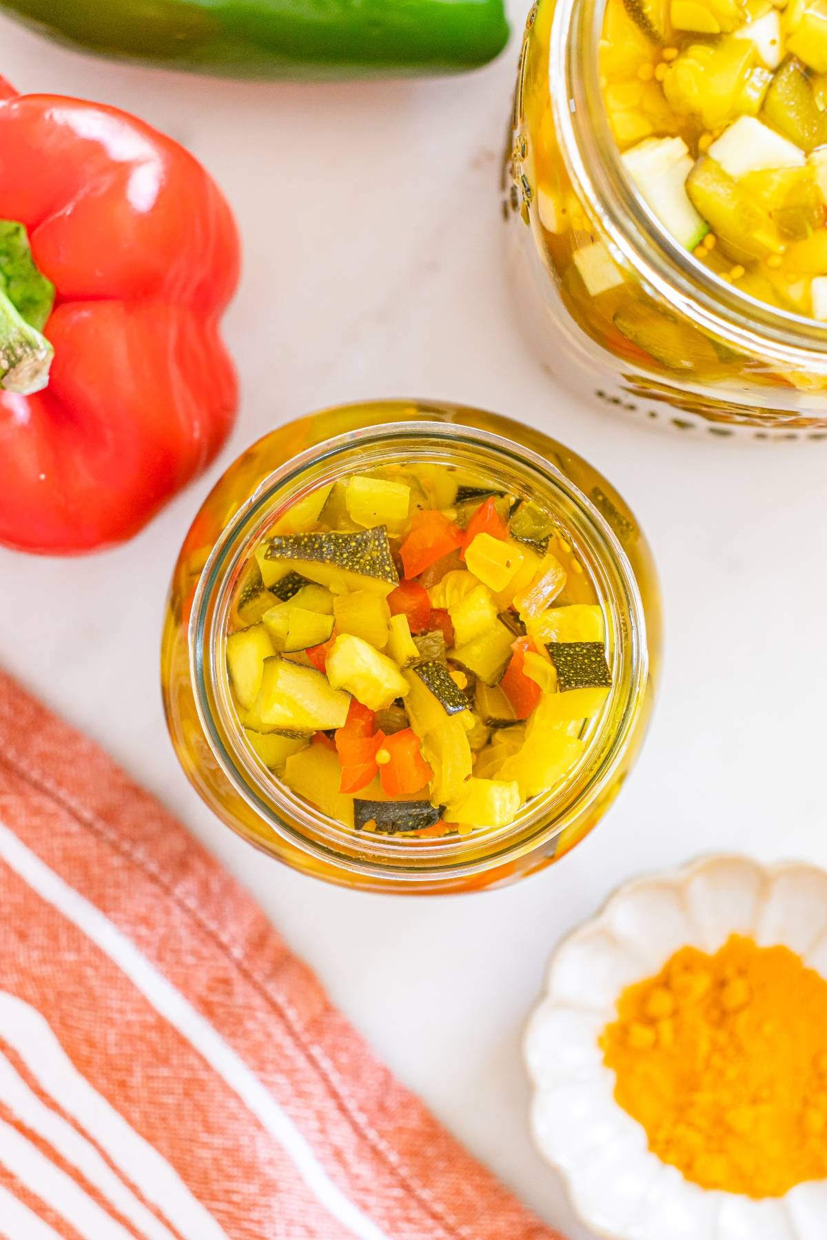 Jar of colorful diced pickled vegetables next to a red bell pepper, spice, and an orange striped towel.