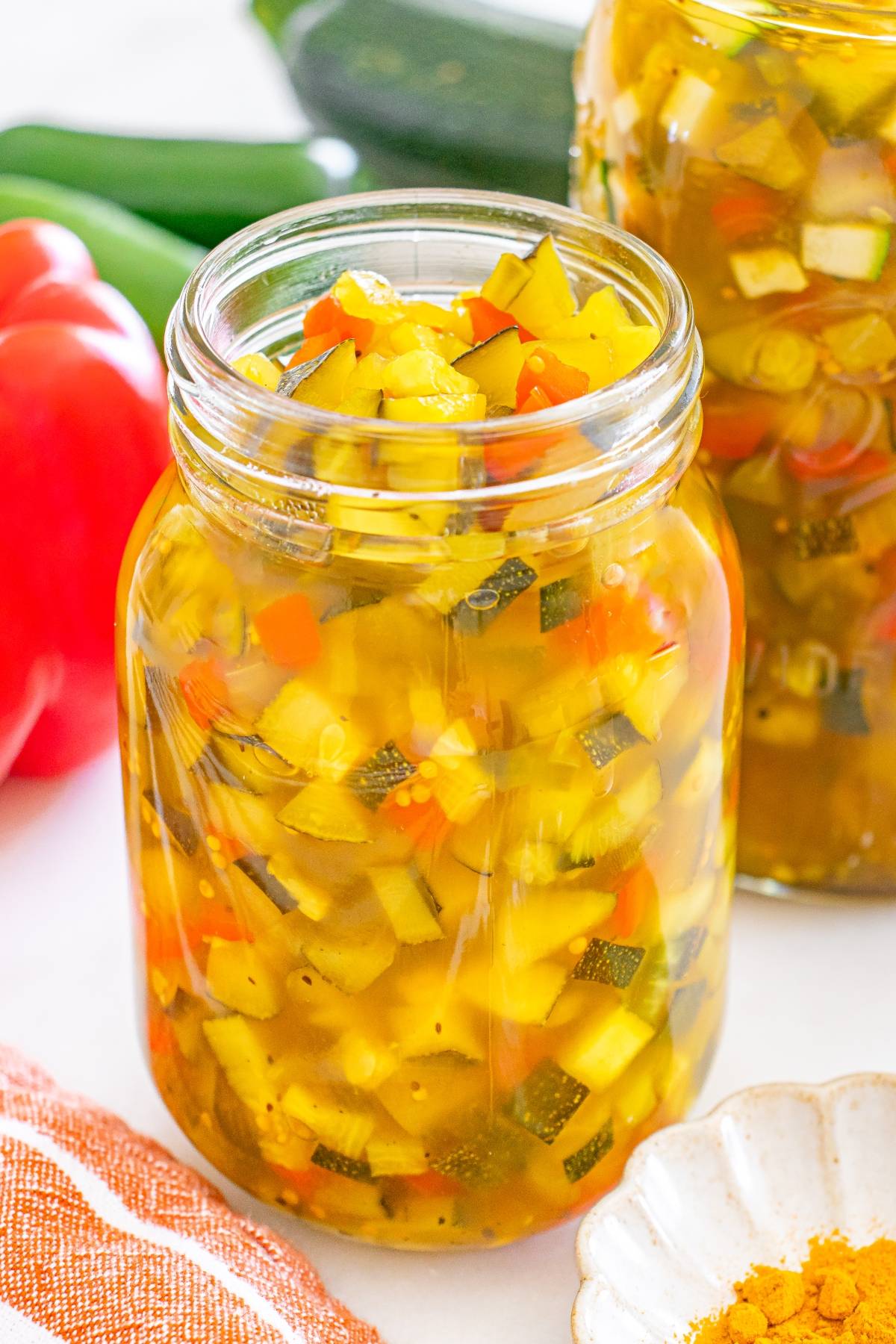 A jar of homemade zucchini relish with diced vegetables in a yellow brine on a white countertop.