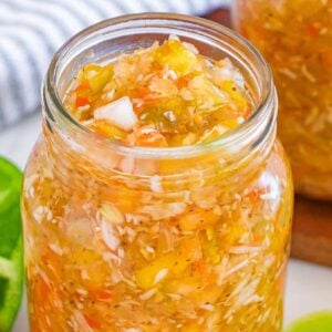 A glass jar filled with tangy homemade chow chow relish, surrounded by fresh peppers and a striped cloth.