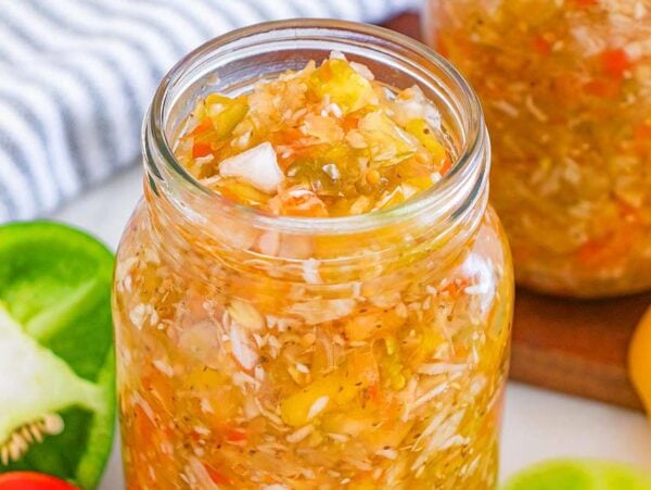 A glass jar filled with tangy homemade chow chow relish, surrounded by fresh peppers and a striped cloth.