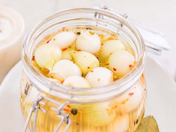 A glass jar of cocktail onions pickled with spices sits next to a container of salt and red pepper flakes.