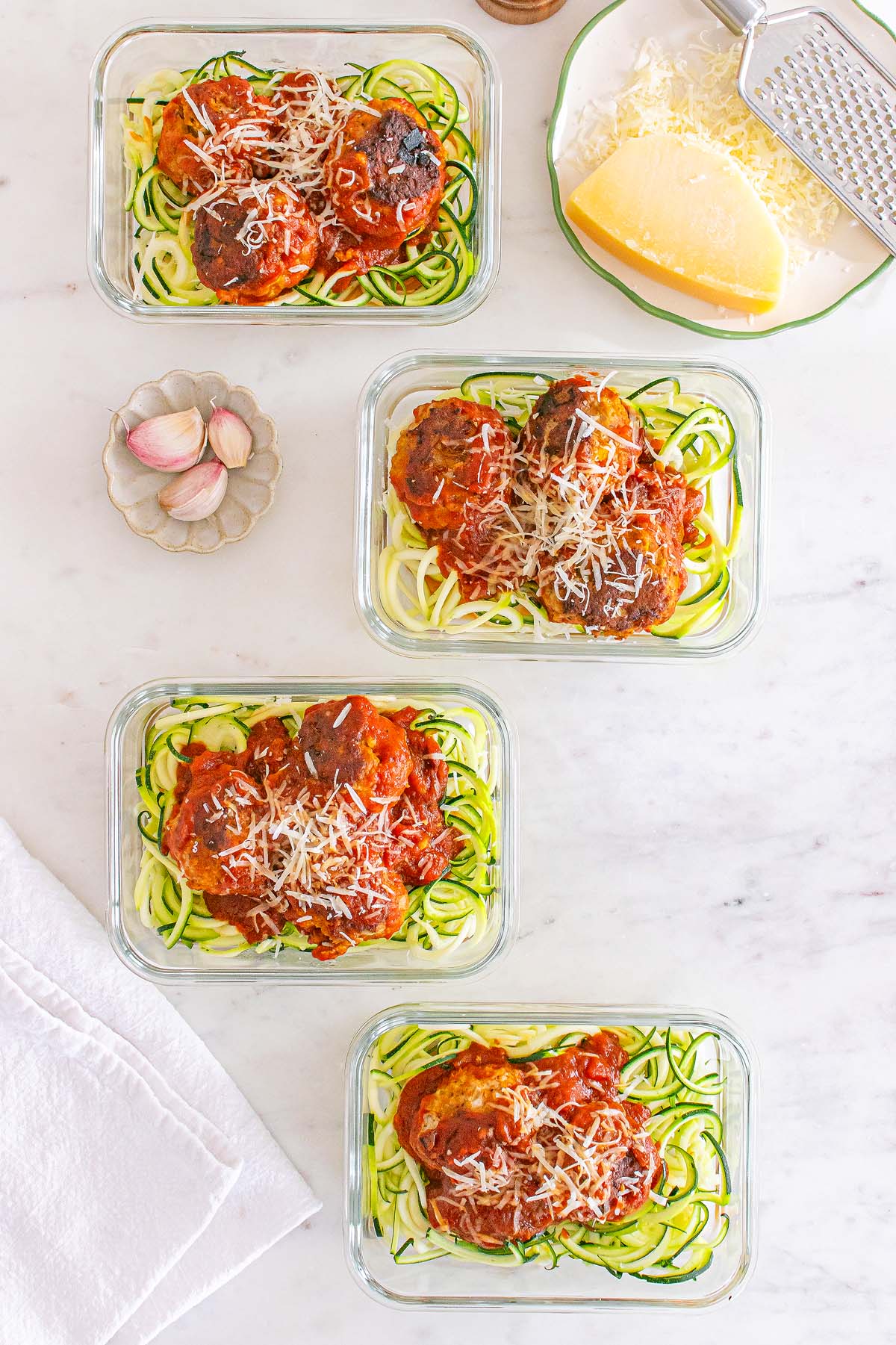 Four glass containers with Meal Prep Turkey Meatballs & Zoodles in tomato sauce, topped with grated cheese, arranged on a marble surface.