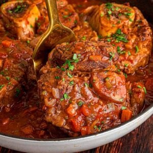 Osso buco braised in rich tomato sauce, garnished with herbs and served in a skillet on a wooden table.