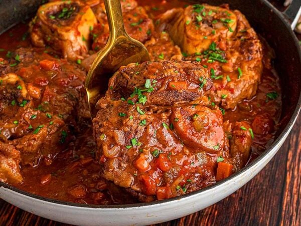 Osso buco braised in rich tomato sauce, garnished with herbs and served in a skillet on a wooden table.