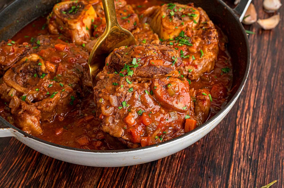 A pan of osso buco braised in tomato sauce, garnished with chopped herbs, with a spoon lifting a tender piece.