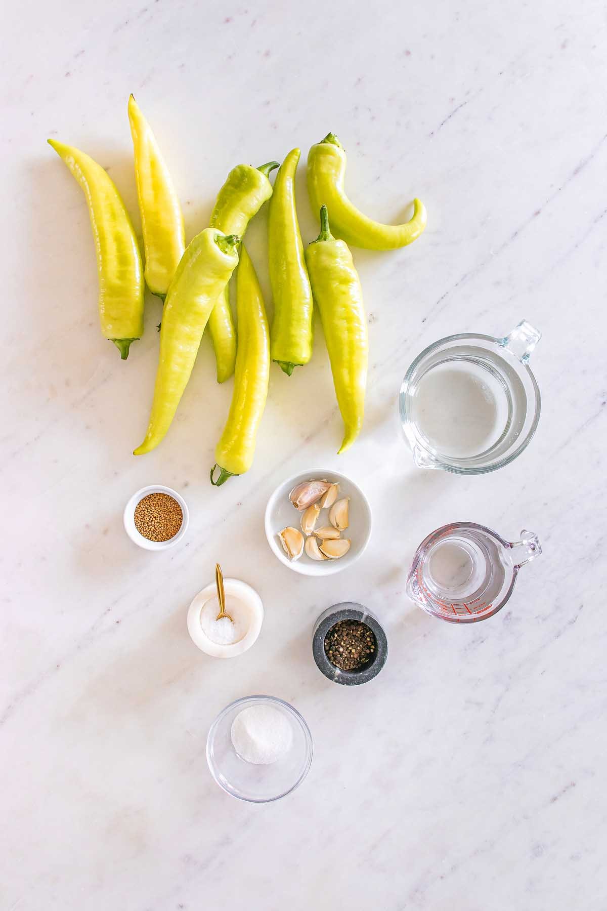 Eight yellow peppers, garlic cloves, seasonings, and measuring cups on a white marble countertop—perfect for making homemade pickled banana pickles.