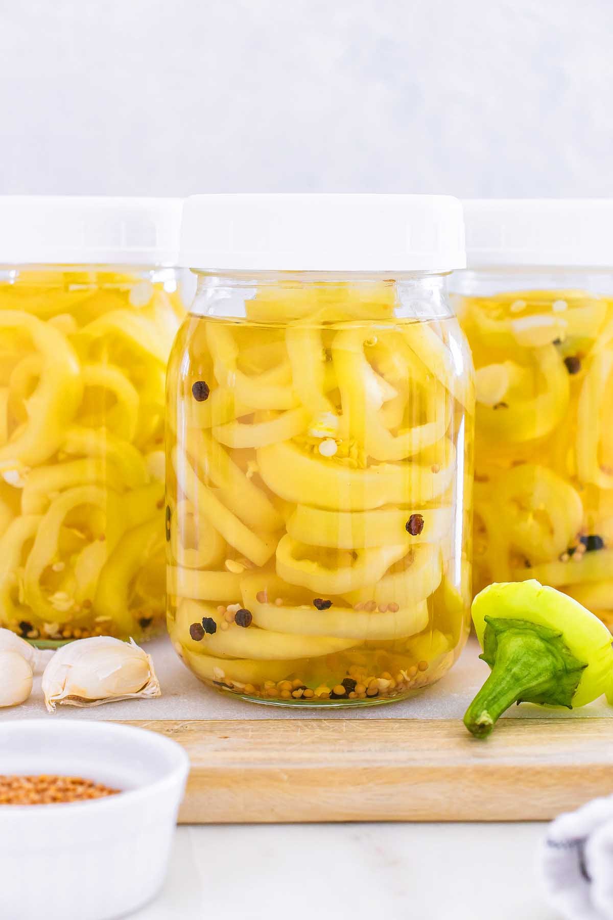 Three jars filled with pickled banana pickles are surrounded by garlic cloves and a fresh pepper on a wooden board.