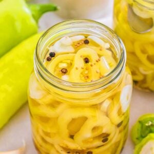 A jar of pickled banana pickles with peppercorns sits next to fresh banana peppers and mustard seeds.