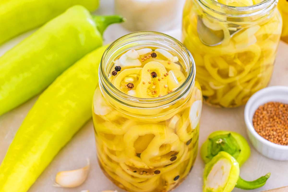 Jars of pickled banana pickles with garlic and spices, surrounded by fresh peppers and seasonings.