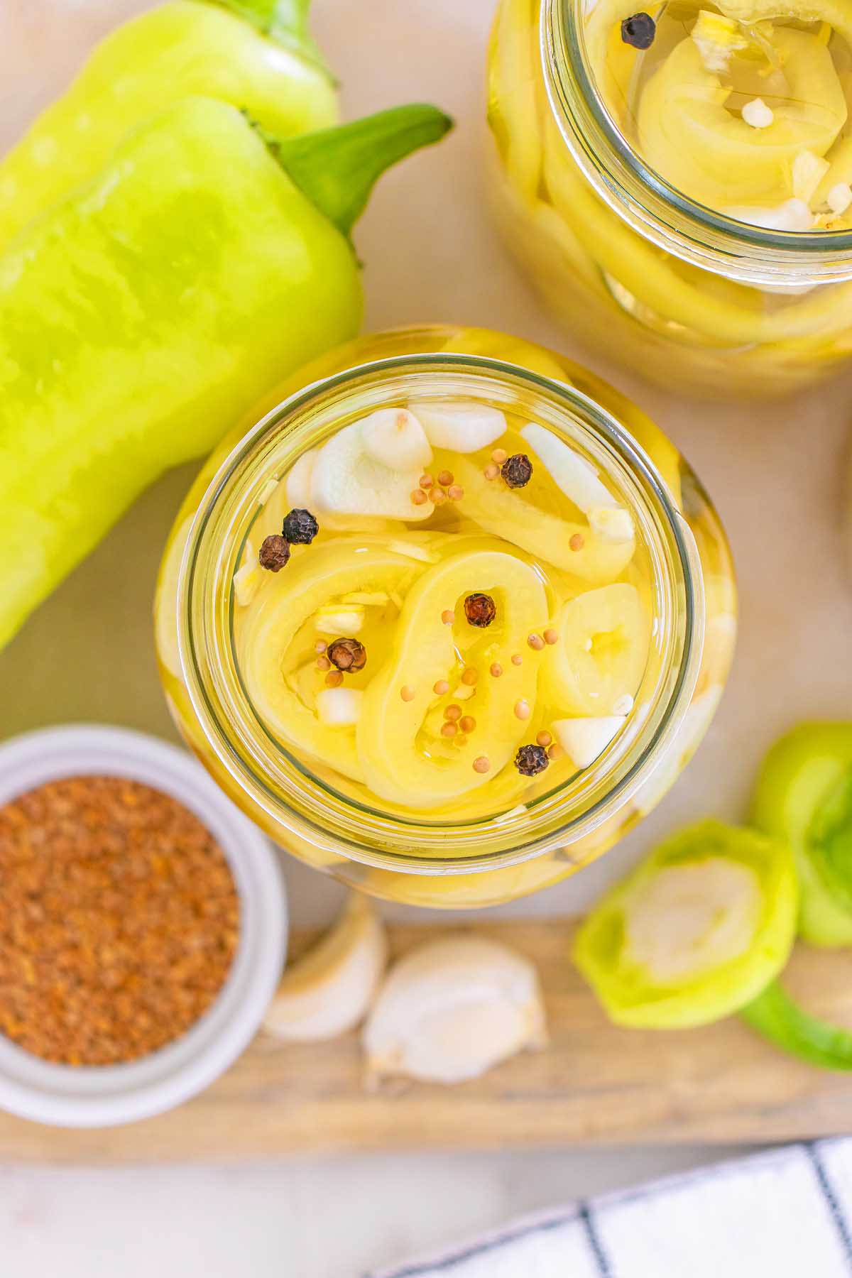 Overhead view of a jar filled with sliced pickled banana pickles, spices, and garlic on a wooden board.