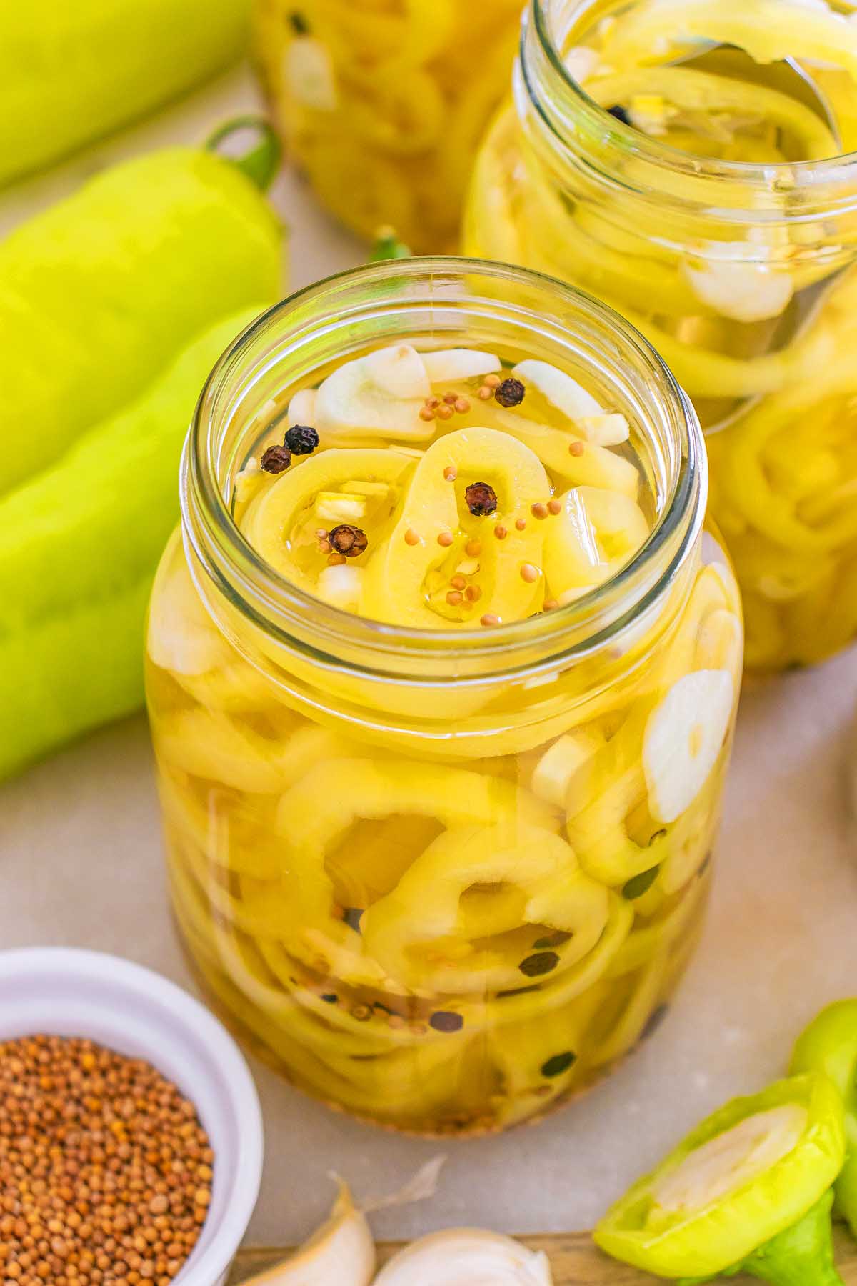 Jar of pickled banana pickles with sliced garlic and spices, surrounded by fresh peppers and mustard seeds.