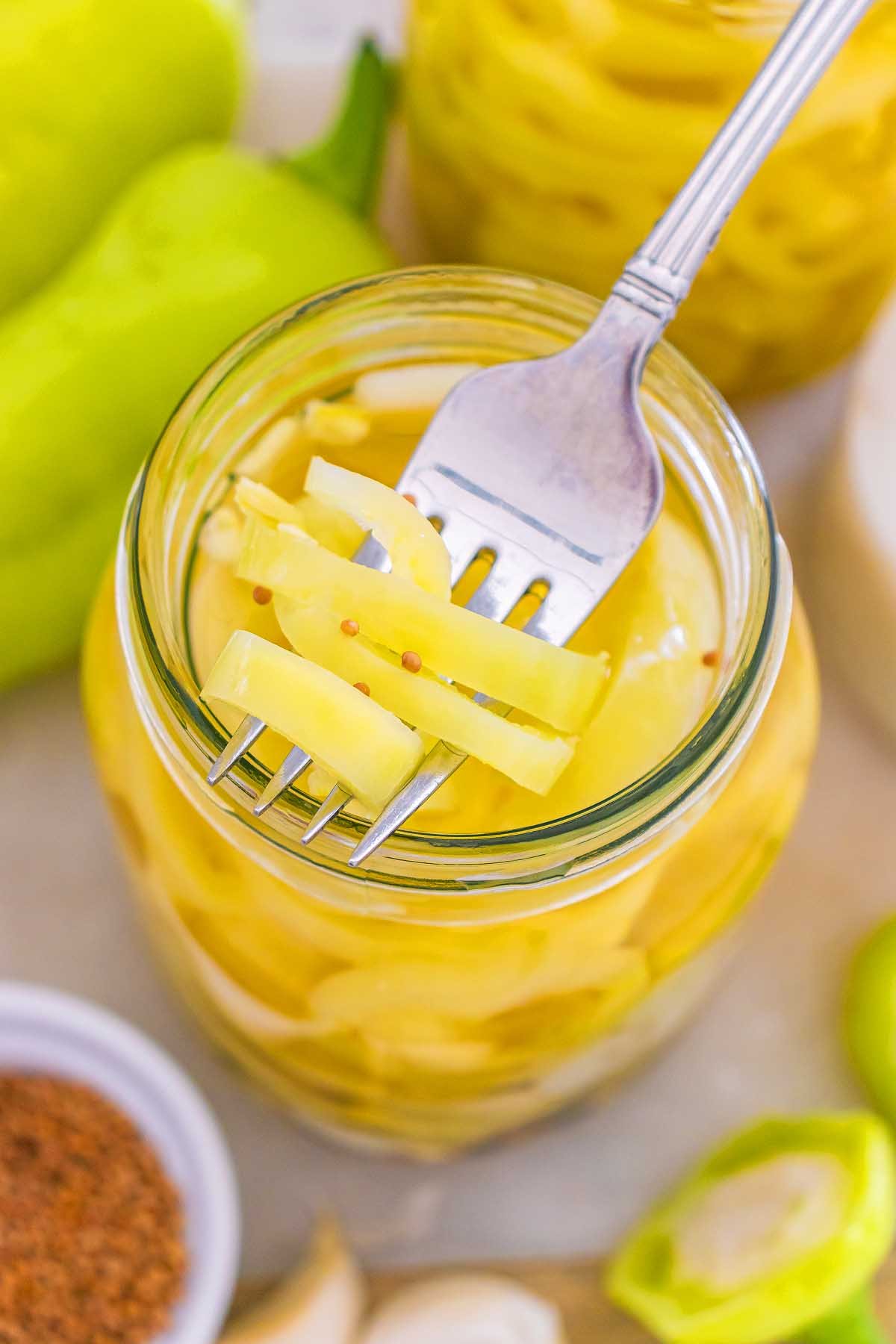 A fork holds sliced pickled banana pickles over an open glass jar filled with more tangy, vibrant pickled peppers.