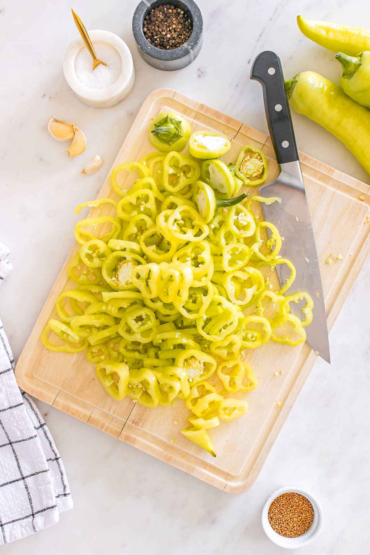 Sliced banana peppers and a knife on a wooden cutting board, with seasonings, whole peppers, and pickled banana pickles nearby.