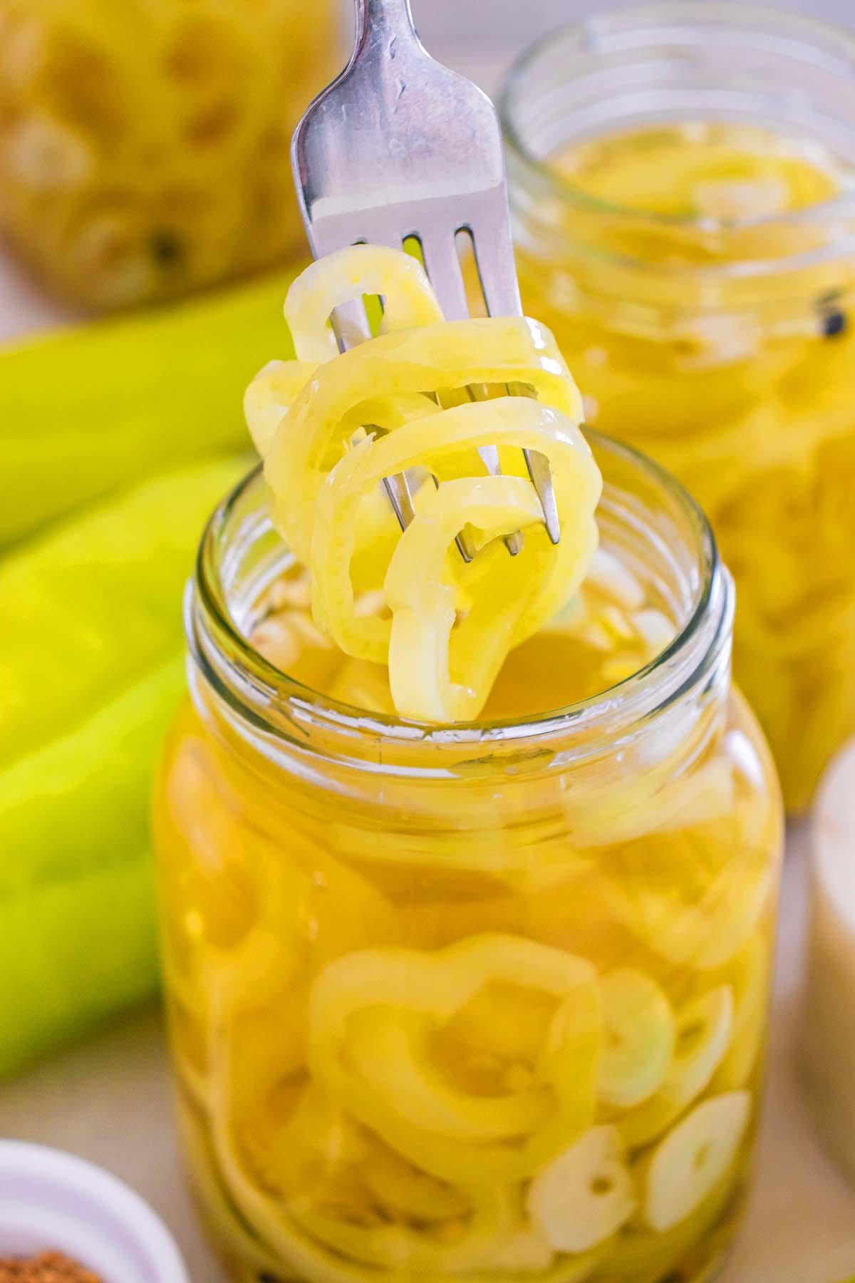 A fork lifts pickled banana pickles from a glass jar filled with brine.