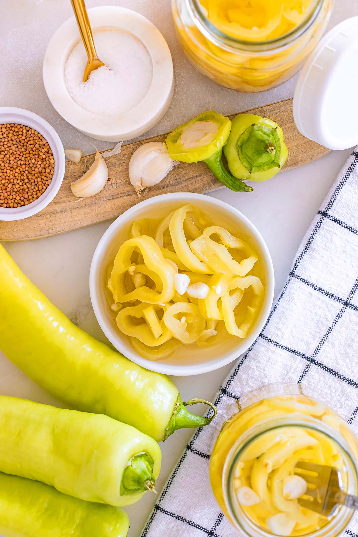 Sliced banana peppers in a bowl with whole peppers, garlic, mustard seeds, salt, and jars on a countertop—perfect for making flavorful pickled banana pickles at home.