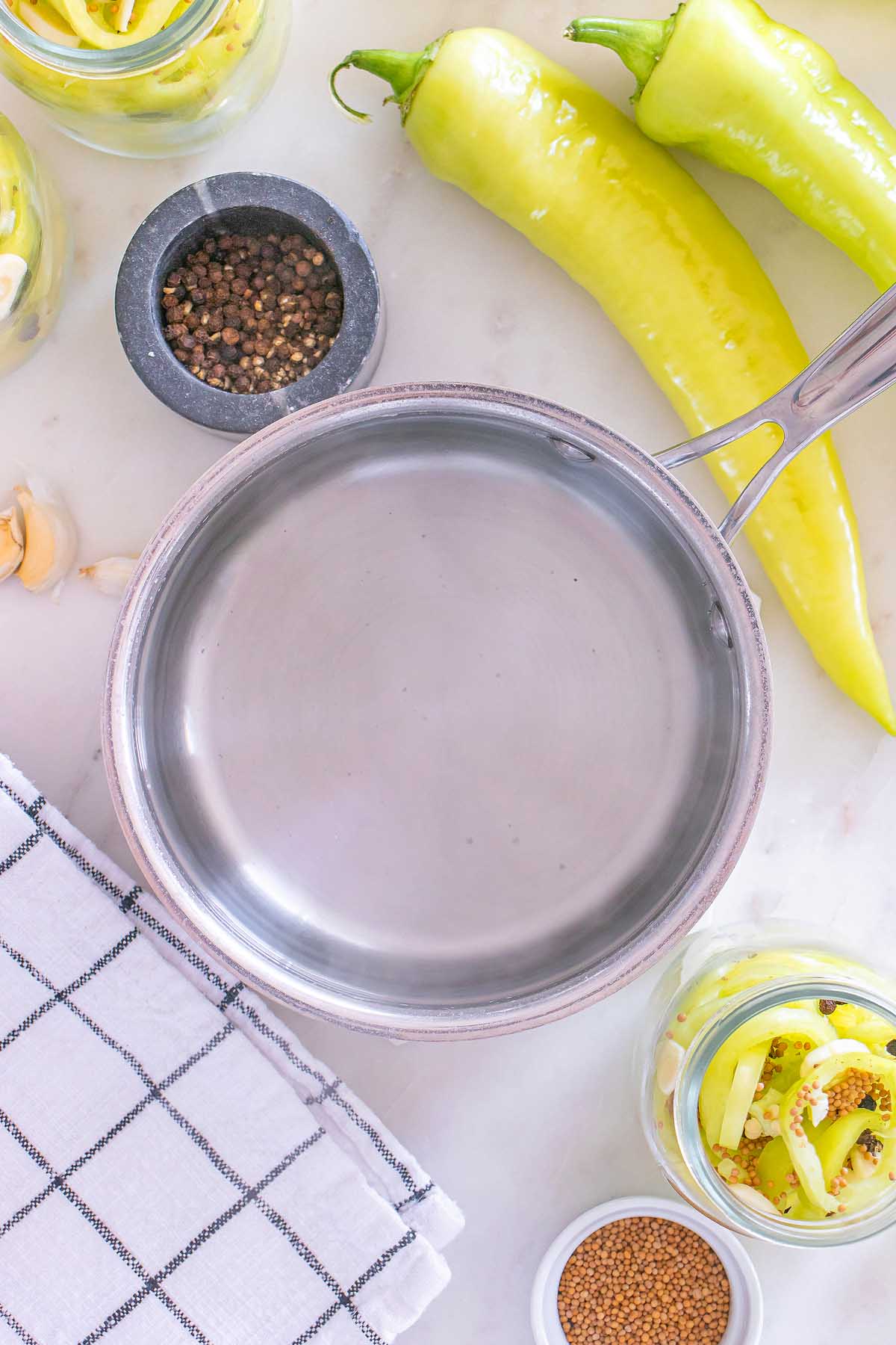 A saucepan, yellow peppers, spices, and a checkered towel on a white surface, ready for making pickled banana pickles.