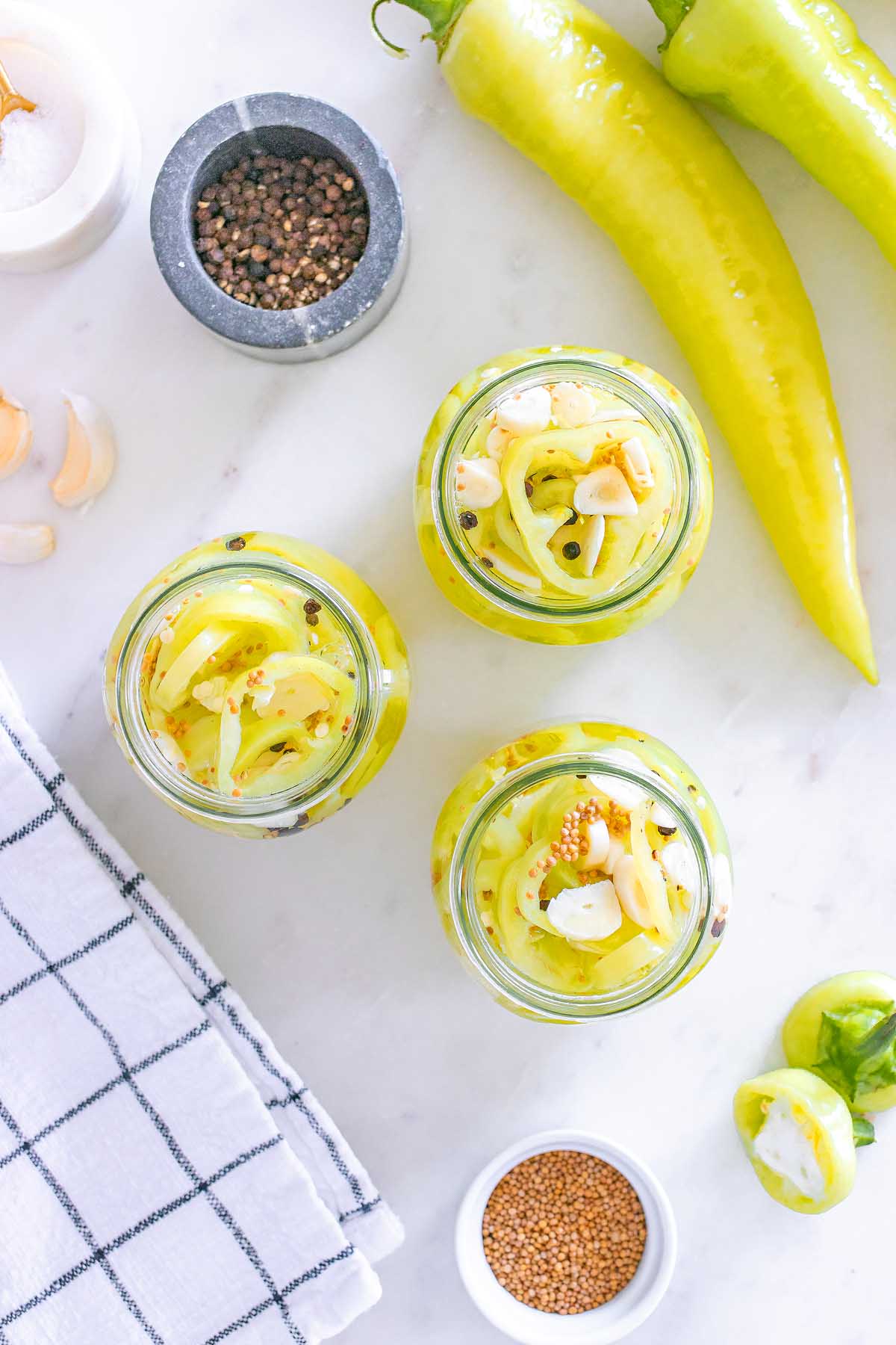 Three jars of pickled banana pickles with garlic and spices sit on a white surface, surrounded by fresh peppers.