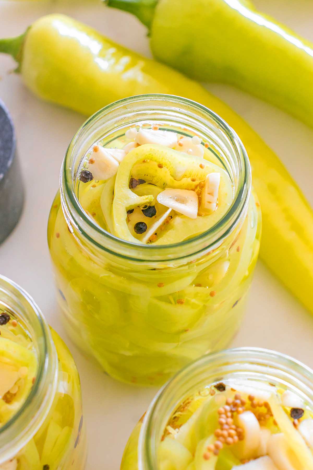 Jar filled with sliced pickled banana pickles, garlic, and spices on a light surface with fresh peppers nearby.