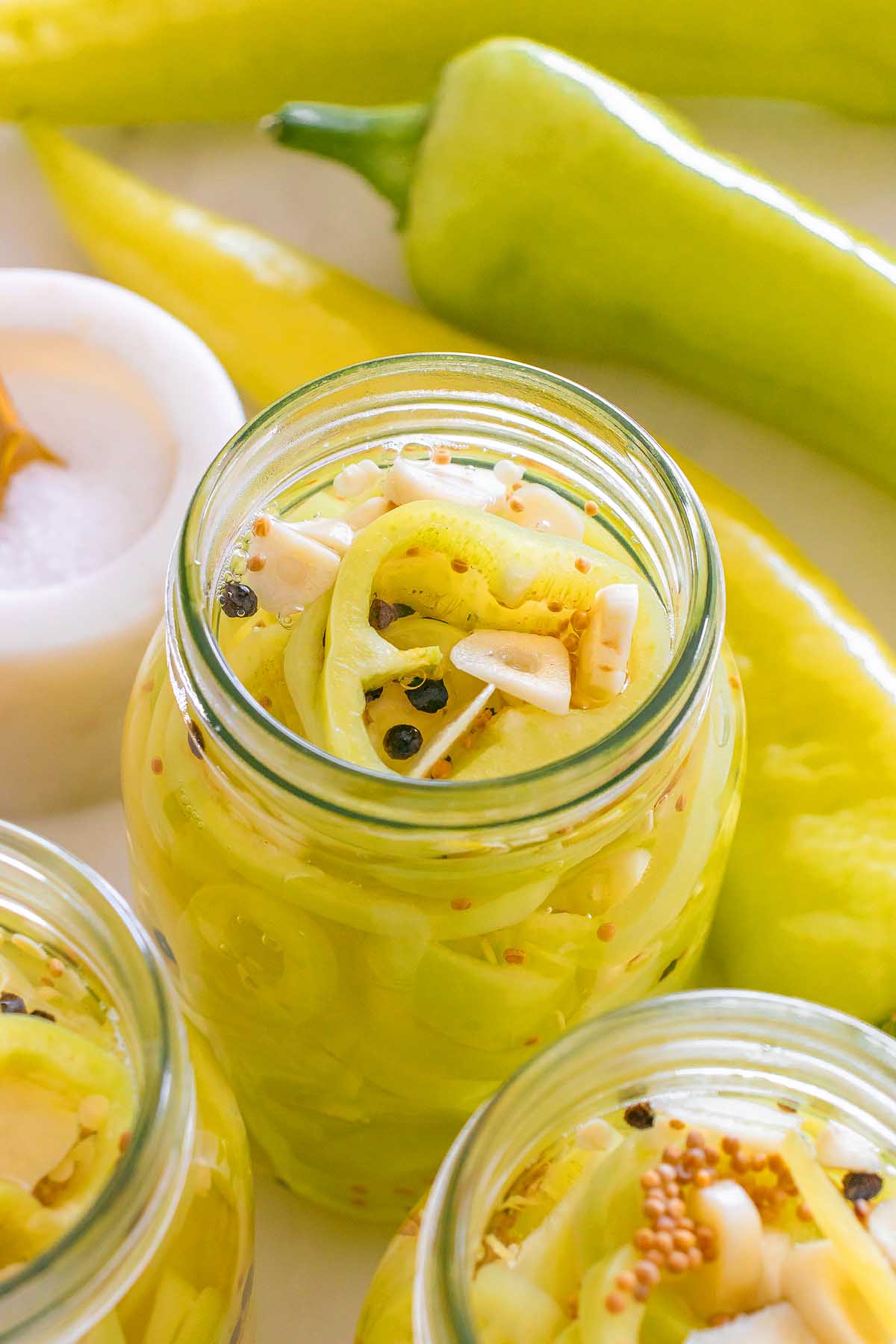 Jar of pickled banana pickles with garlic, mustard seeds, and peppercorns, surrounded by fresh peppers.