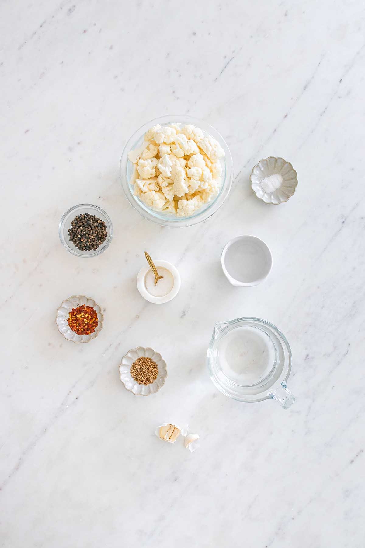 Pickled cauliflower florets, spices, garlic, and water arranged in small bowls on a white marble surface.