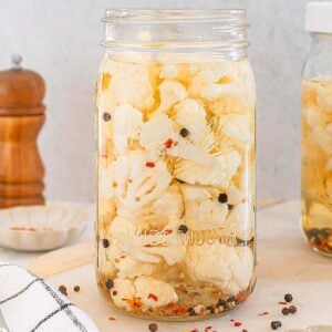 Two jars of pickled cauliflower with spices sit on a kitchen counter, accompanied by a knife and a pepper grinder, ready to enhance your next meal.