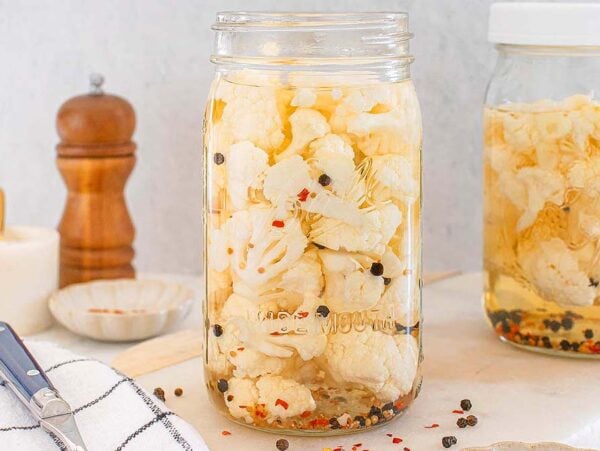 Two jars of pickled cauliflower with spices sit on a kitchen counter, accompanied by a knife and a pepper grinder, ready to enhance your next meal.