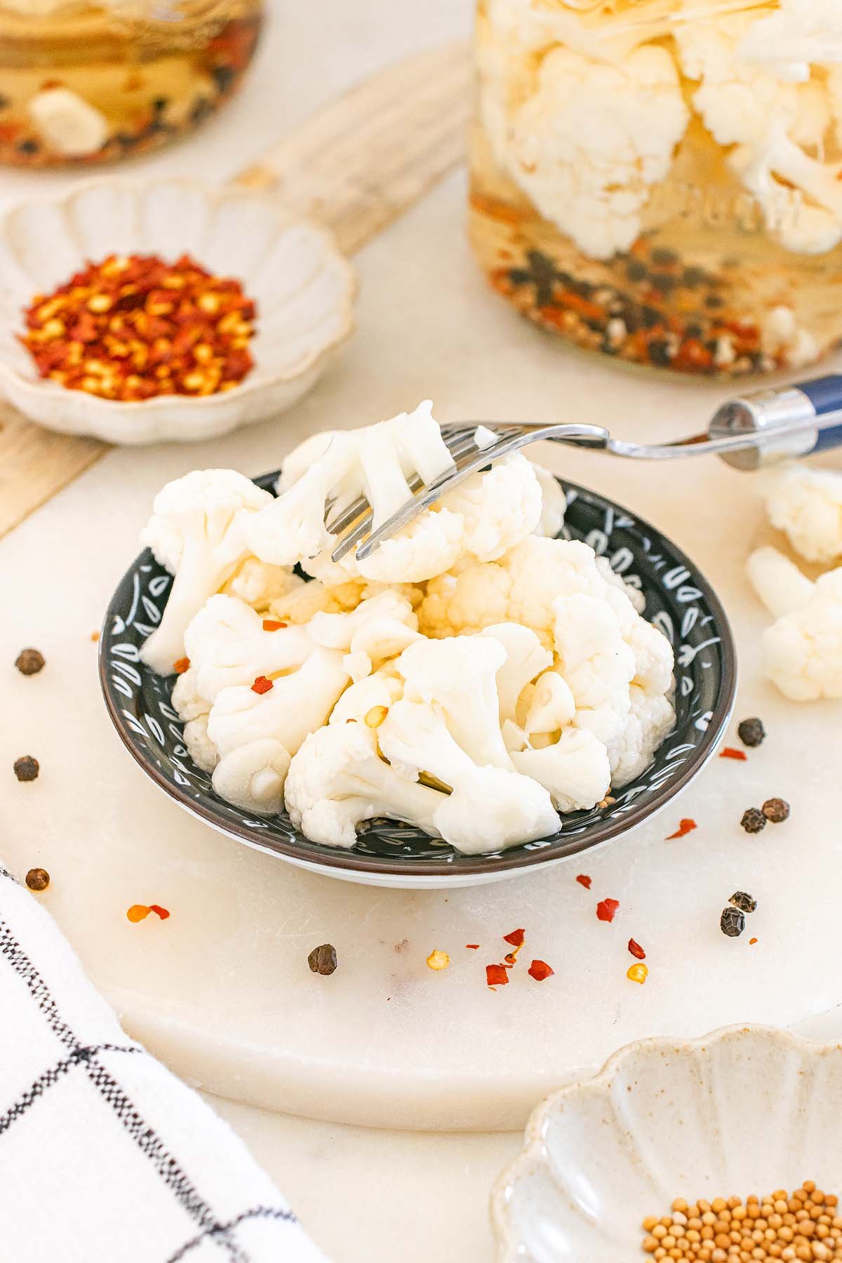 A fork holds tangy pickled cauliflower in a small bowl, surrounded by spices and jars filled with more pickled cauliflower.