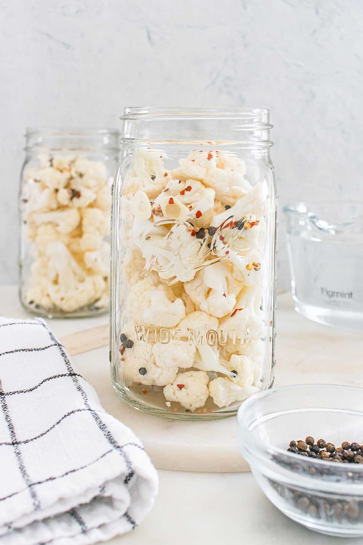 Glass jar filled with pickled cauliflower, spices, and herbs sits on a counter near a towel and other jars.