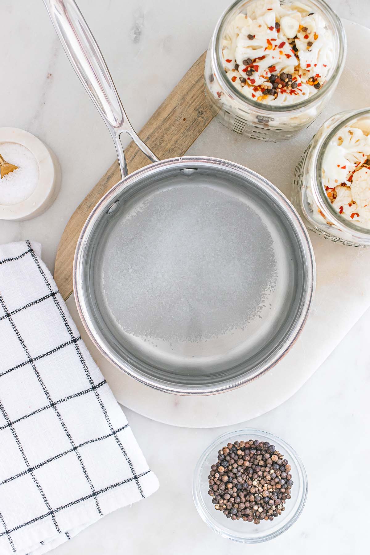 A saucepan with water sits on a counter beside black peppercorns, salt, and two jars of pickled vegetables, including pickled cauliflower.
