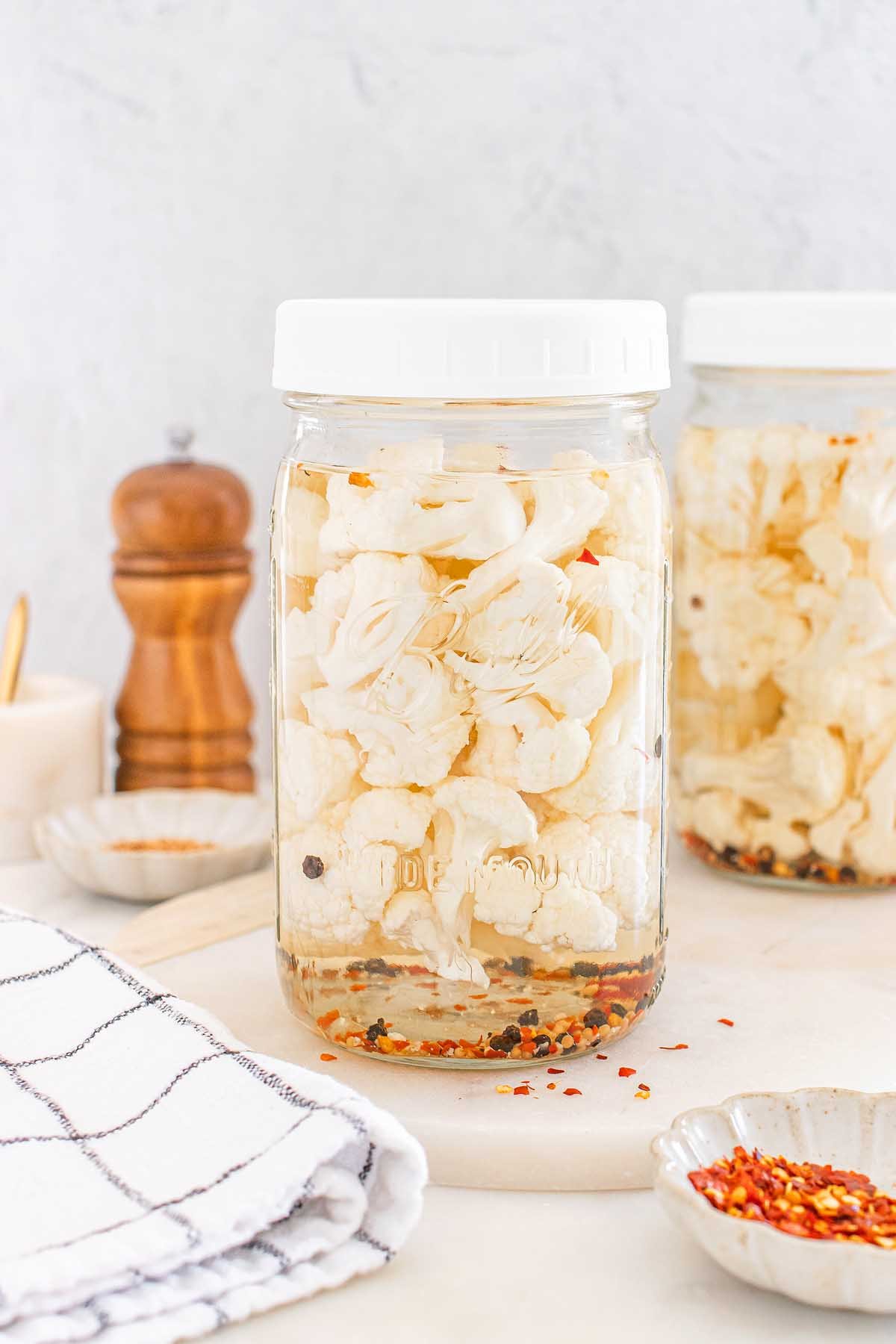 A glass jar filled with pickled cauliflower florets, spices, and brine sits on a white countertop.