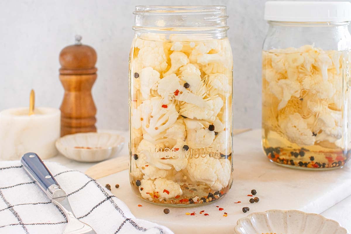 Two jars of pickled cauliflower with peppercorns sit beside a pepper grinder and kitchen towel, ready to enjoy.