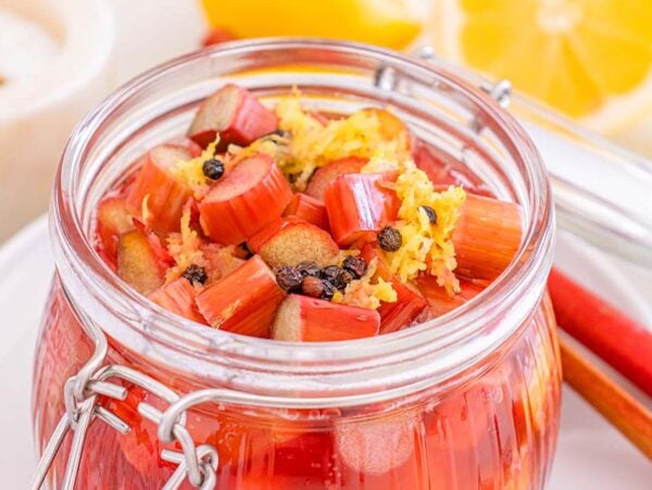 Glass jar filled with pickled rhubarb, lemon zest, and spices, with lemon slices and rhubarb stalks arranged nearby.