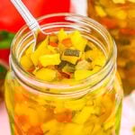 A jar of colorful homemade zucchini relish with a spoon inside, surrounded by fresh vegetables and spices.