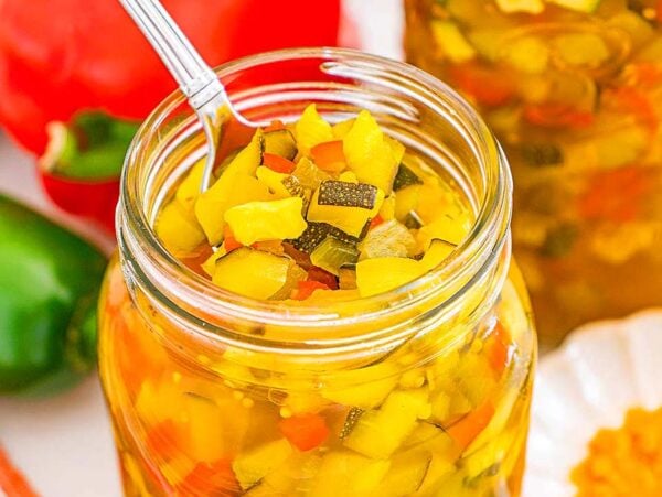 A jar of colorful homemade zucchini relish with a spoon inside, surrounded by fresh vegetables and spices.