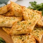 Slices of air fryer garlic bread on a wooden board with a small bowl of herb butter and fresh parsley nearby.