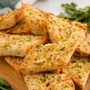 Slices of air fryer garlic bread on a wooden board with a small bowl of herb butter and fresh parsley nearby.