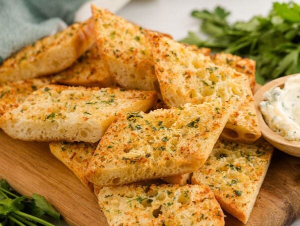 Slices of air fryer garlic bread on a wooden board with a small bowl of herb butter and fresh parsley nearby.