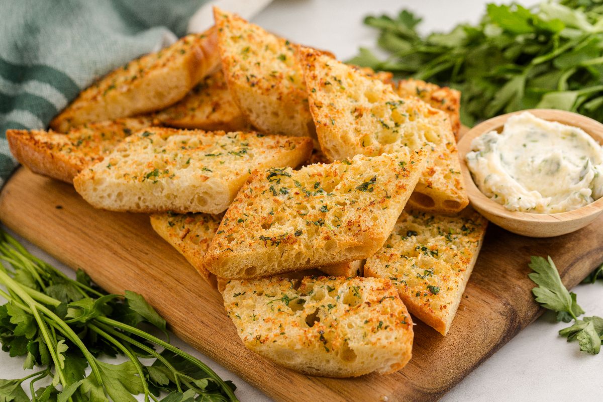 Slices of air fryer garlic bread on a wooden board with herbs and a bowl of garlic butter nearby.