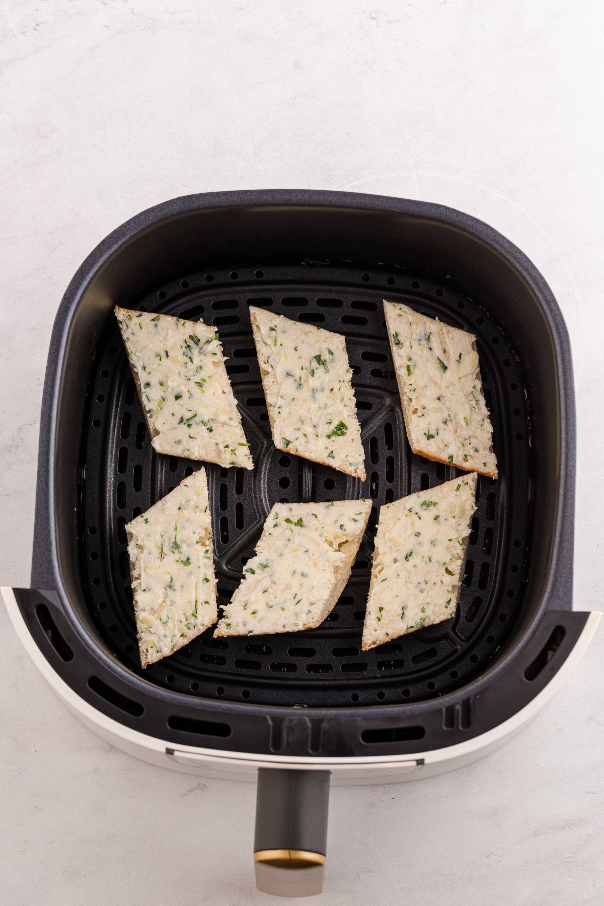 Six uncooked diamond-shaped flatbreads with herbs in an air fryer basket on a white surface.